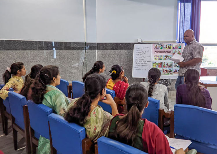 Male staff member conducts a presentation in front of a group of women, facing away from the foreground. The man is pointing to a board with a number of images on it. 