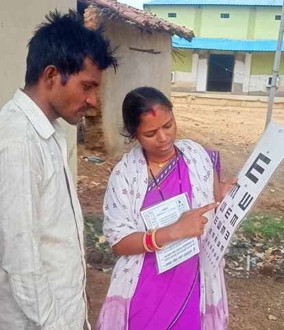 Female health worker wearing a purple sari stands beside a male patient wearing a white shirt, as she conducts a vision test. 