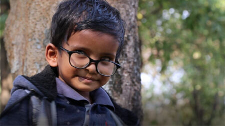 Alok poses for a photo outside, standing in a wooded area in front of a tree. He wears a warm jacket and a pair of black eyeglasses.