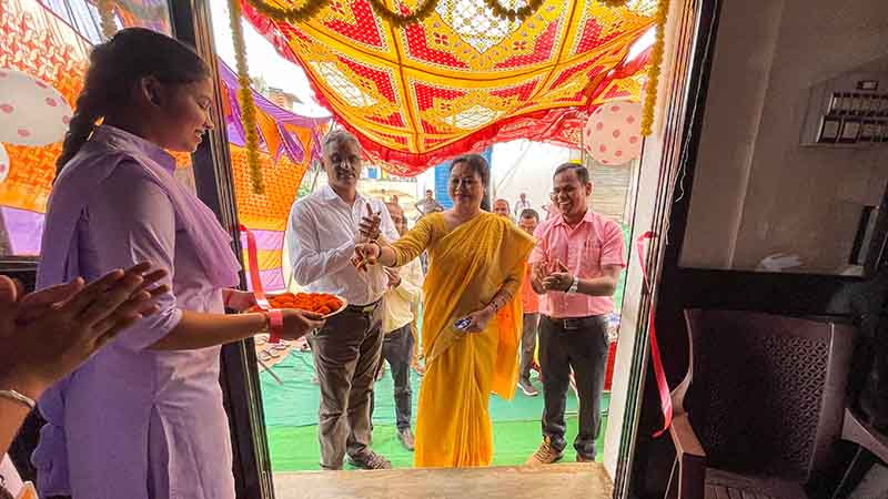 Woman in a purple sari in the foreground, holds a copper plate full of marigold flowers, in the background stand a woman with men standing on either side, having just cut a ribbon.
