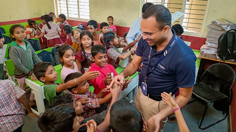 A smiling man shakes the hands of children who crowd around him in a classroom.