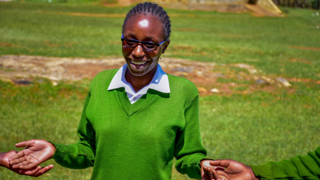 A teenaged girl wearing glasses is standing in a field, smiling.