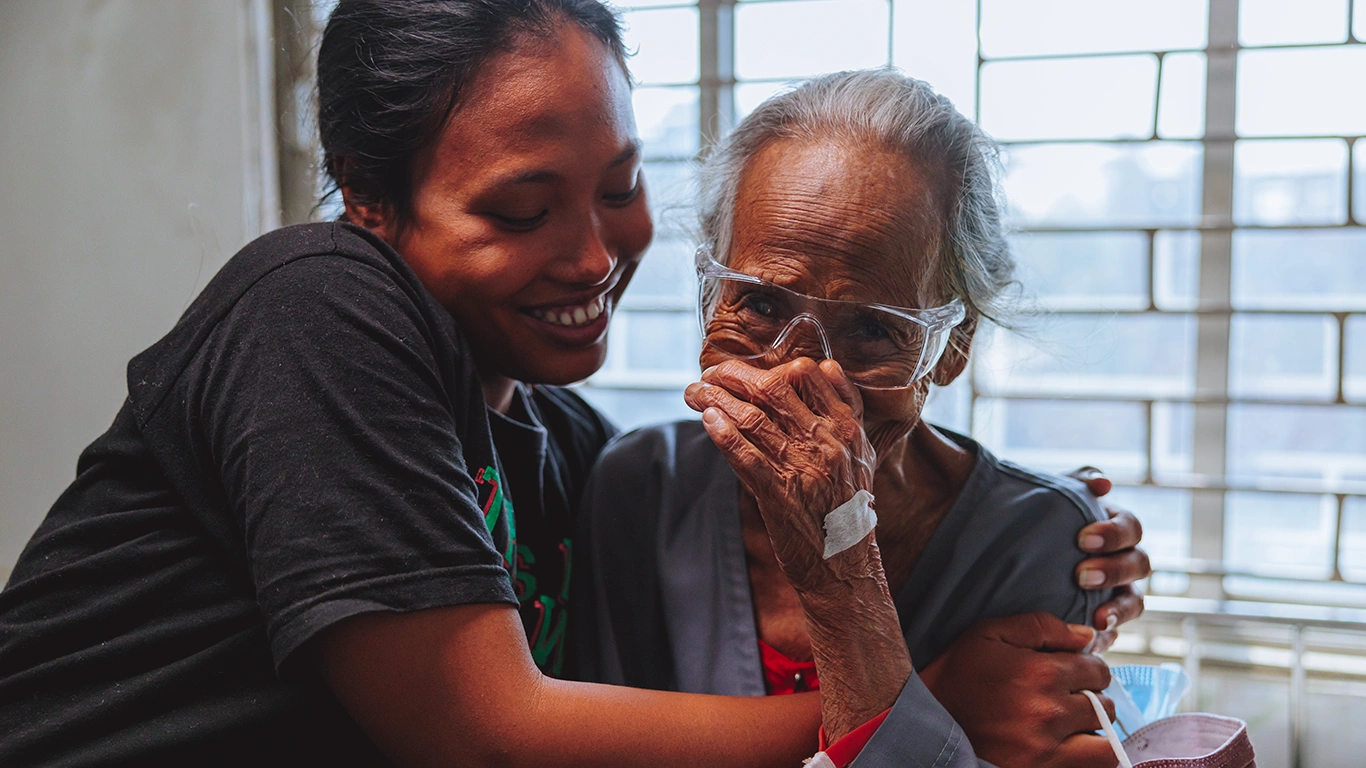 A young woman hugs an older woman, who is wearing protective eyeglasses and appears to be wiping away tears. They both smile.