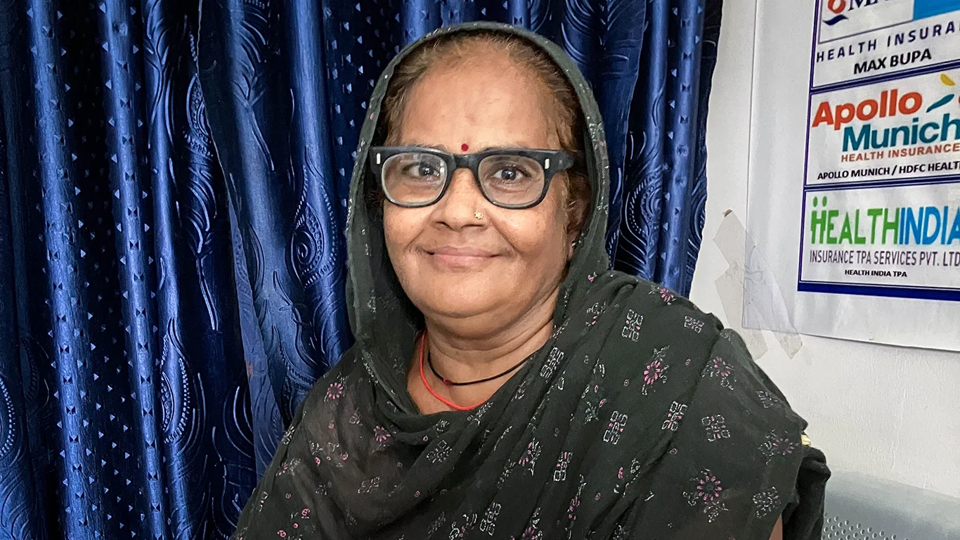 A woman sits in a clinic setting, smiling at the camera. She wears black eyeglasses.