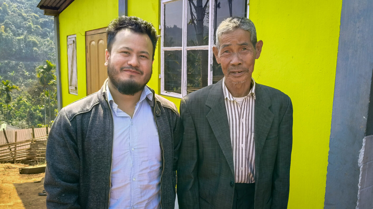 A young man poses, smiling, with an older man outside a yellow house.